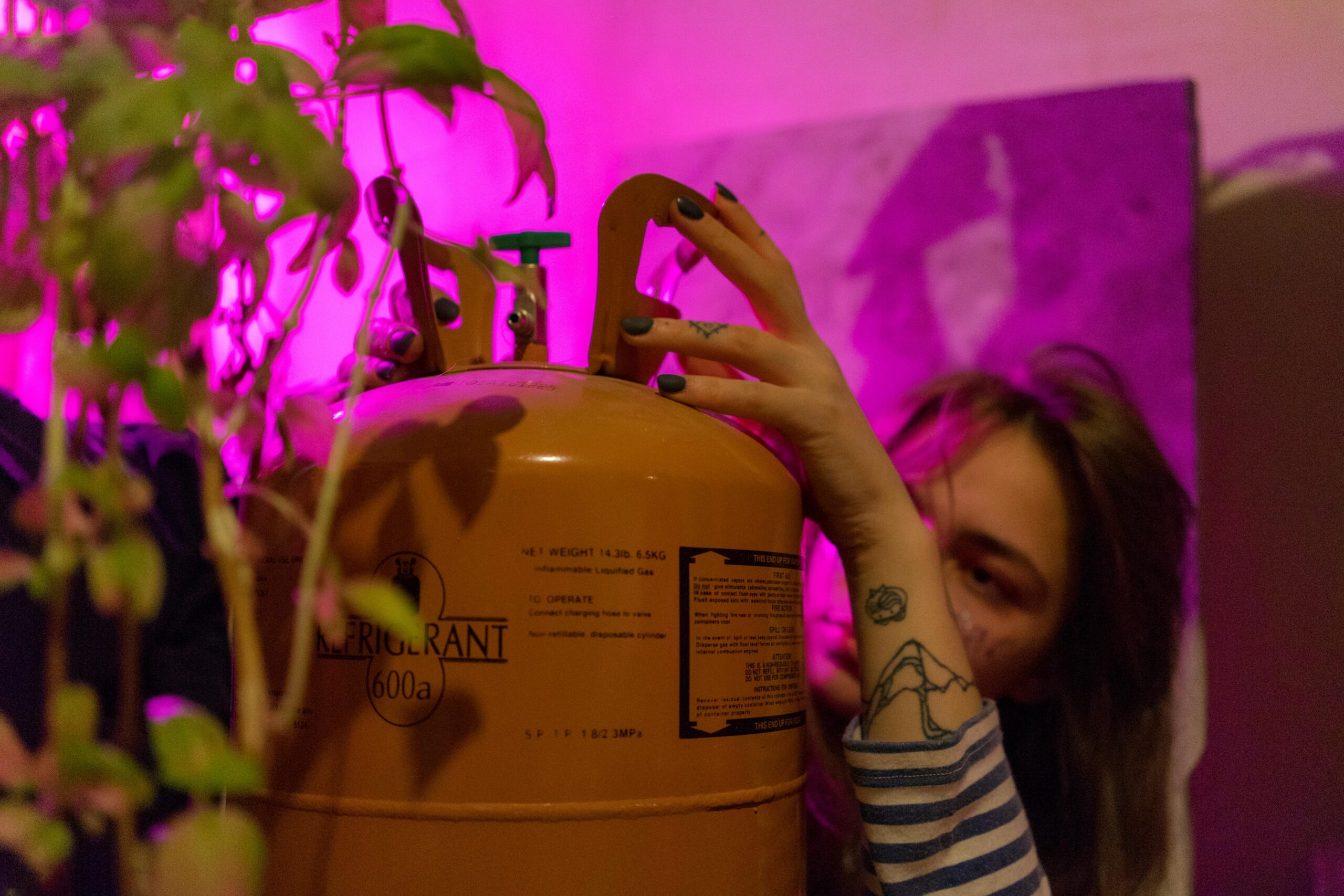 A young woman in a modern indoor setting with a tattoo, holding a refrigerant tank under vivid violet neon light.